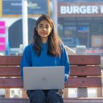 Teenage girl sitting on a bench using a laptop in an urban outdoor setting.