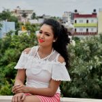A cheerful woman sitting outdoors in a white lace top against a cityscape backdrop.